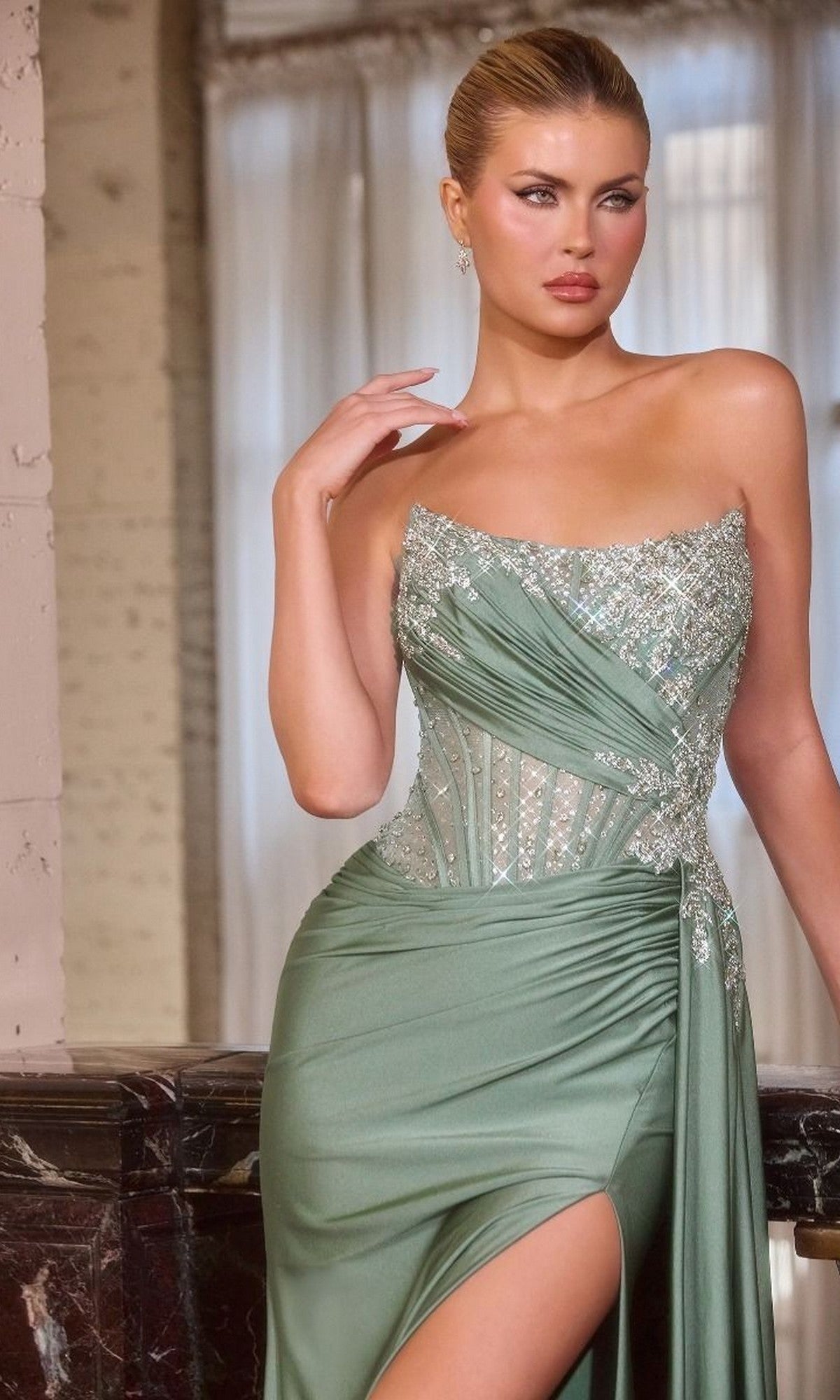 A woman stands indoors near a window, wearing the Ladivine CD689 long prom dress—a strapless satin gown in light green with silver embellishments, a sheer corset bodice, and a thigh-high slit.